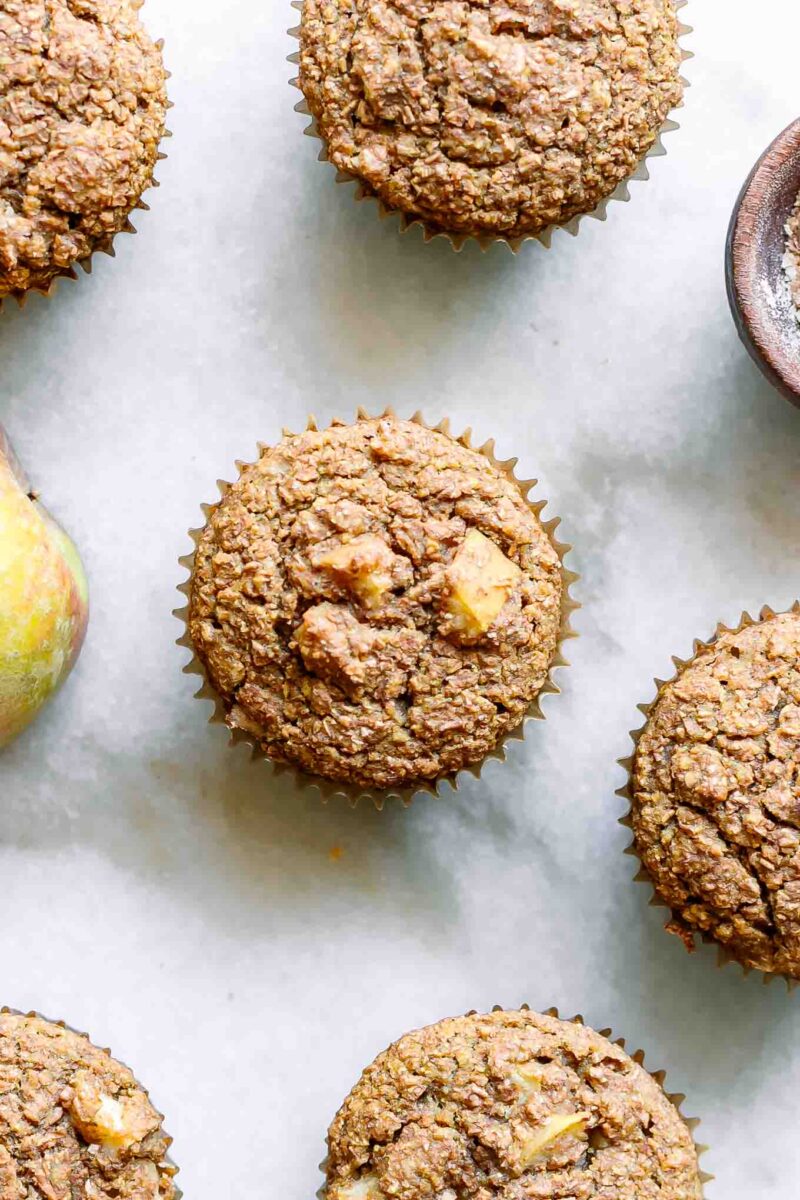 apple bran muffins on a table with a whole apple and bowl of wheat bran