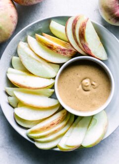 a bowl with peanut butter and apple slices on a white table with apples