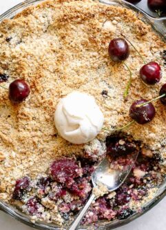 a baking dish with a vegan cherry crisp topped with fresh cherries and a scoop of ice cream on a white table