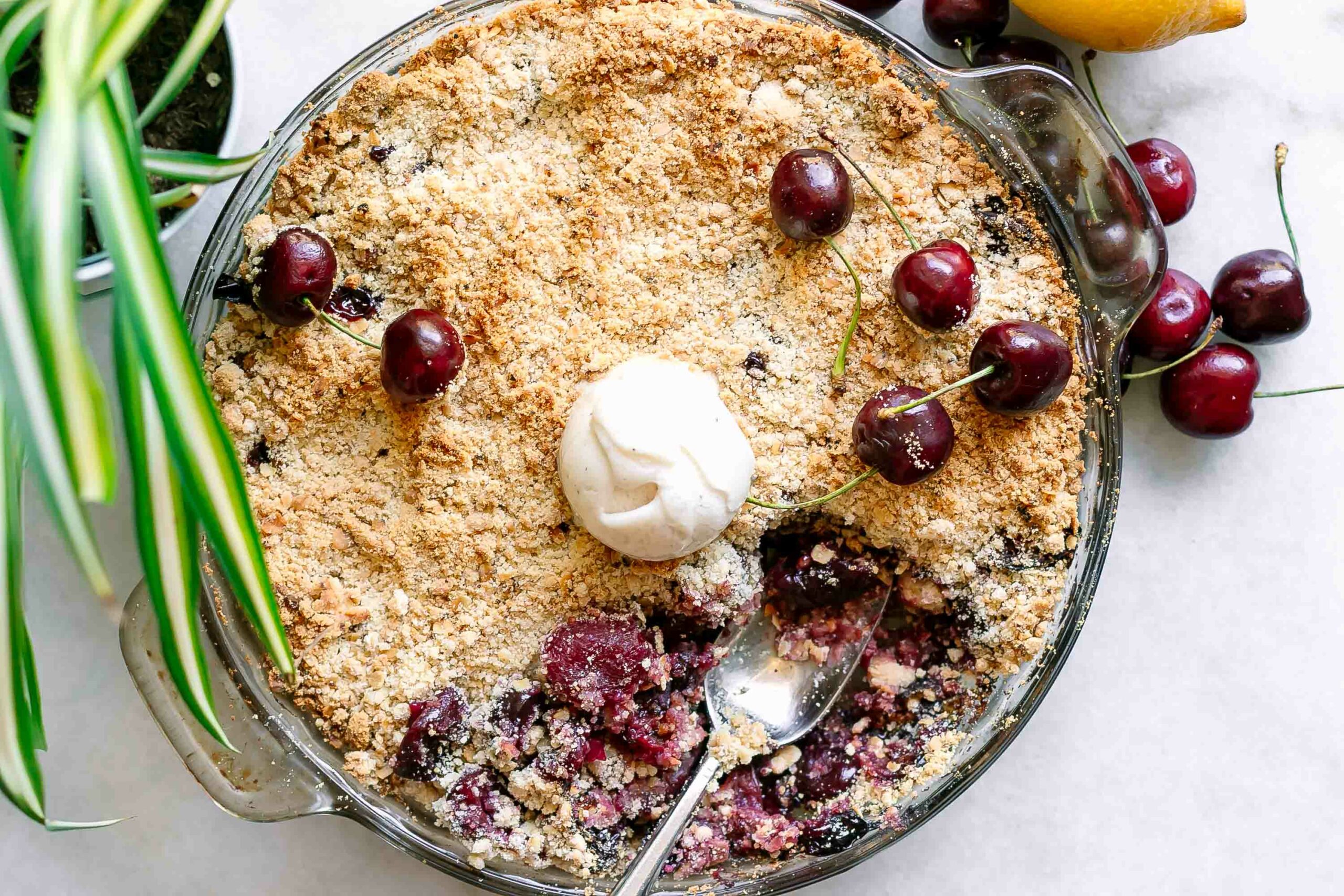 a baking dish with a vegan cherry crisp topped with fresh cherries and a scoop of ice cream on a white table