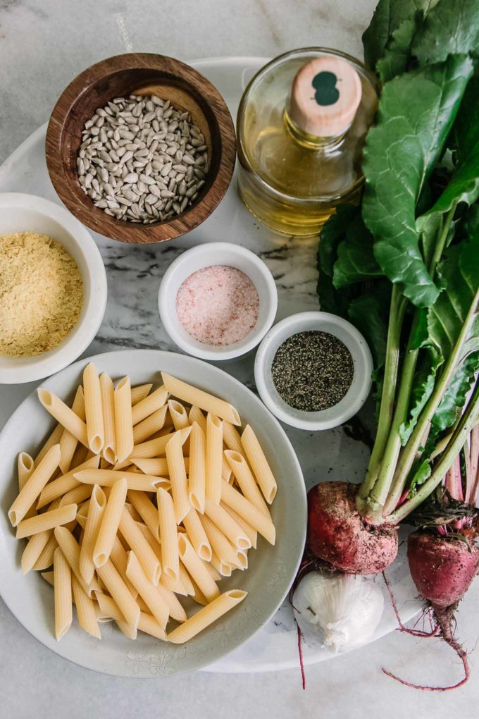 beets with greens and bowls of dry penne pasta, sunflower seeds, nutritional yeast, oil, salt, and pepper on a white table