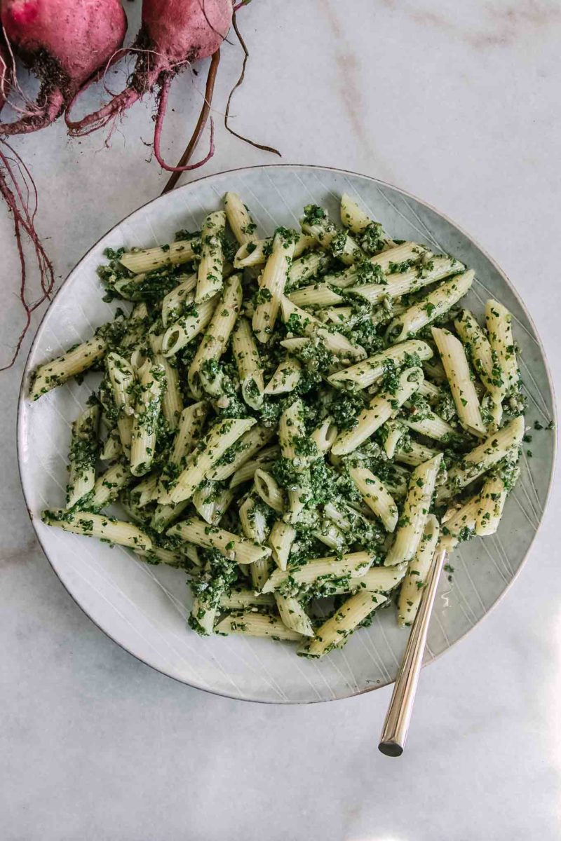 penne pasta noodles with pesto made of beet leaves on a white plate with a gold fork and red beets in the background