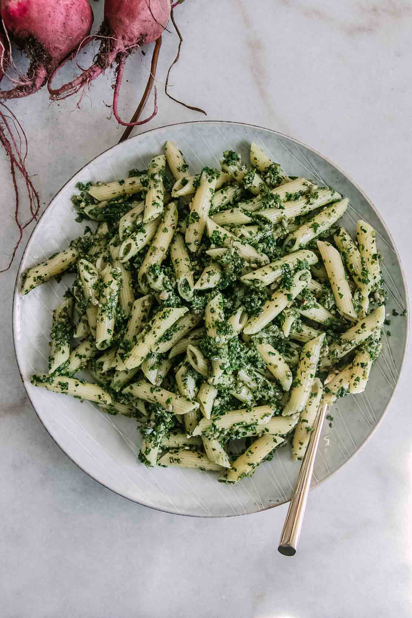 penne pasta noodles with pesto made of beet leaves on a white plate with a gold fork and red beets in the background