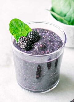 a glass of blackberry spinach smoothie with a spinach garnish on a white table