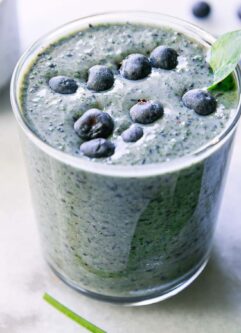a spinach blueberry smoothie in a glass with blueberries and a spinach leaf as garnish on a white table