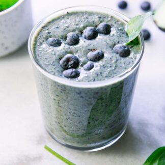 a spinach blueberry smoothie in a glass with blueberries and a spinach leaf as garnish on a white table