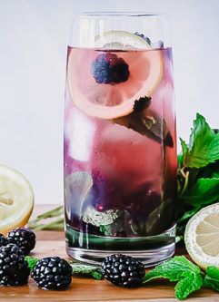 A glass of boozy blackberry lemonade on a cutting board with fresh fruit.