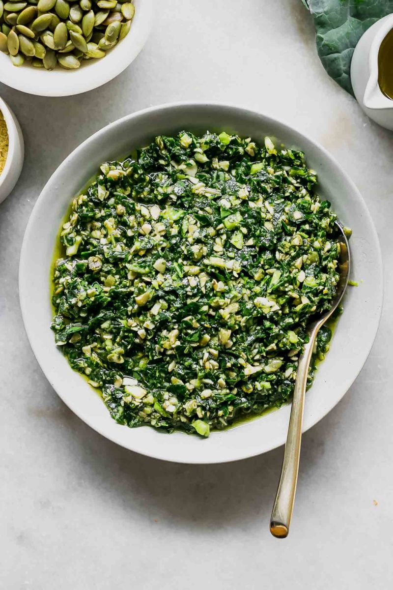 a bowl of pesto with brussels sprouts leaves on a white table with a gold spoon