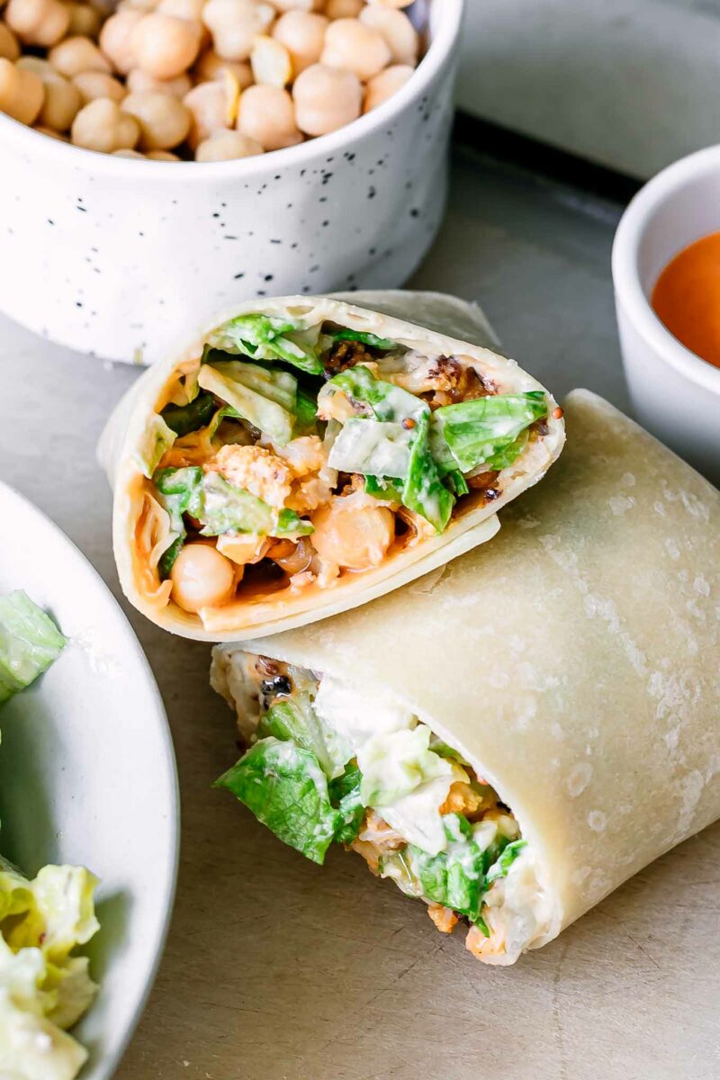 two buffalo caesar wrap halves on a table with a bowl of chickpeas and a small bowl of buffalo sauce