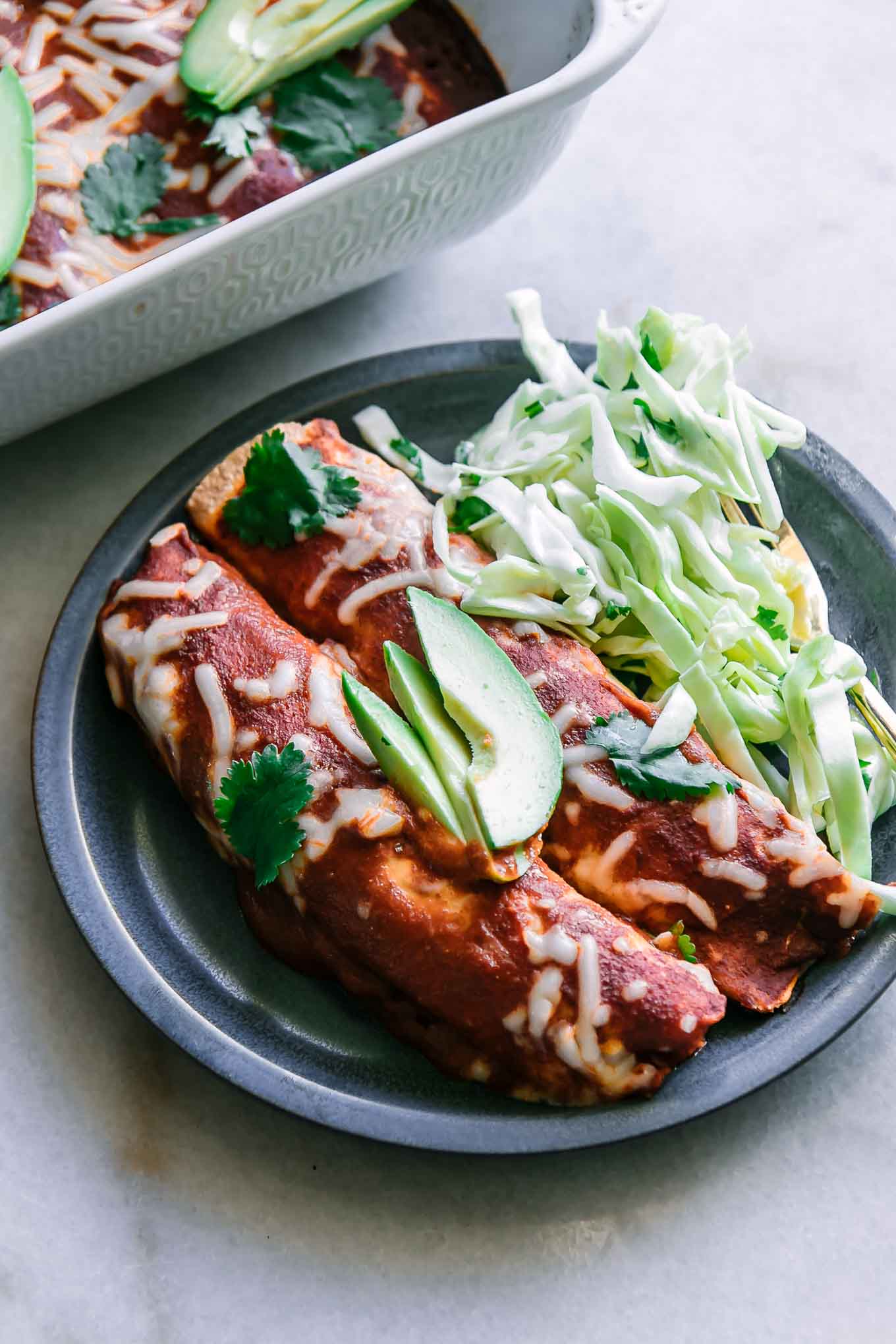 a blue plate with black bean butternut squash enchiladas on a white table with a baking dish filled with enchiladas