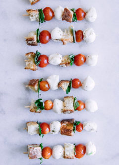Skewers with cherry tomatoes, mozzarella, basil, and bread crumbs on a stick on a white table.