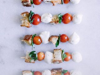 Skewers with cherry tomatoes, mozzarella, basil, and bread crumbs on a stick on a white table.