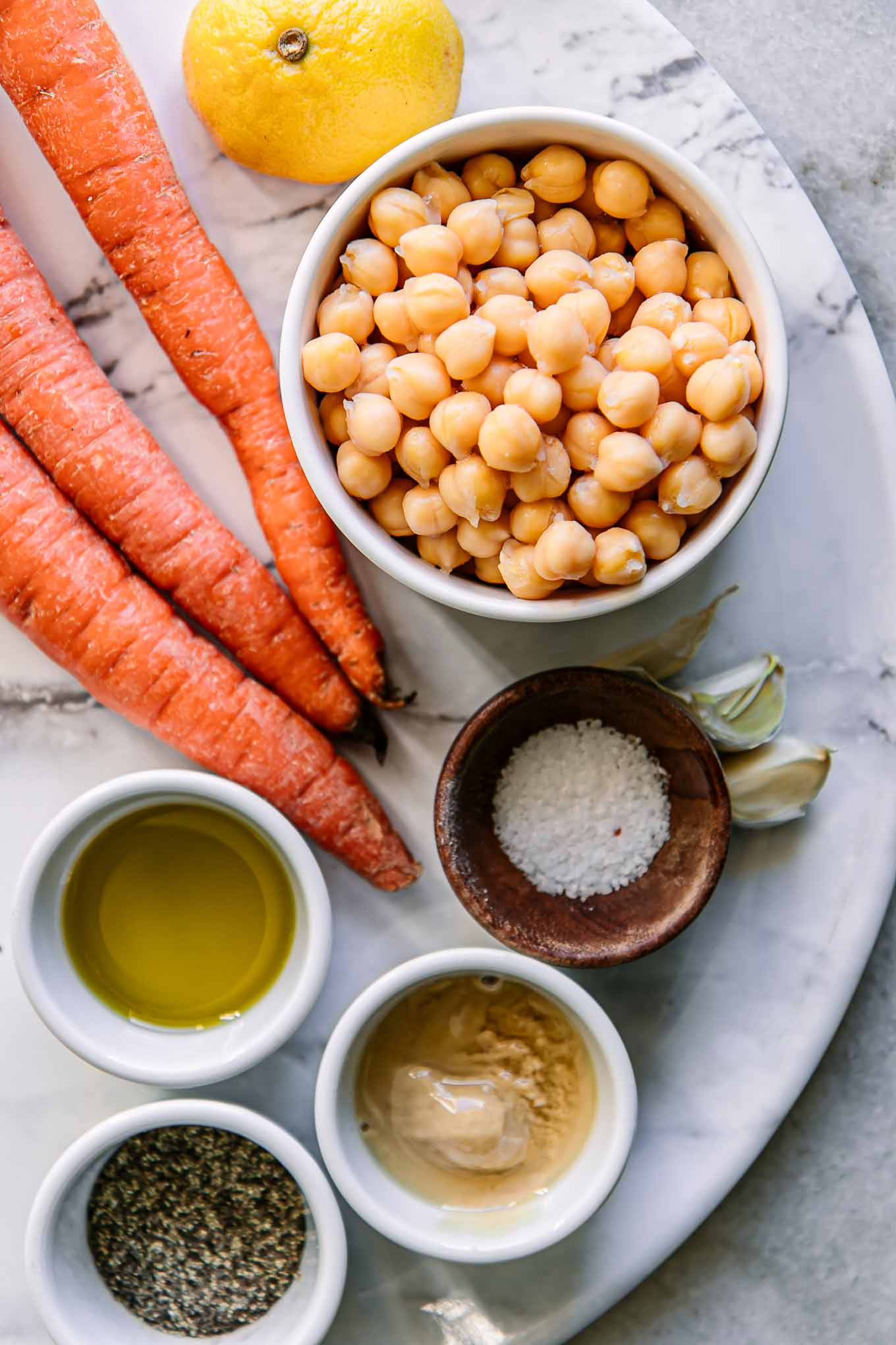 bowls of chickpeas, carrots, tahini, oil, salt, pepper, and lemon on a white table