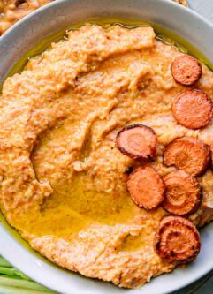 a bowl of carrot hummus dip on a plate with vegetables for dipping