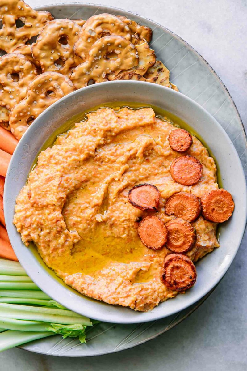 a bowl of orange carrot hummus dip on a plate with celery, carrots, and pretzels