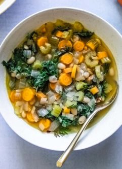 a bowl of vegetable soup with leafy carrot greens in a white bowl on a blue table