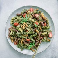 pesto pasta with cherry tomatoes on a white plate on a white table with carrots and a bowl of tomatoes