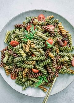 pesto pasta with cherry tomatoes on a white plate on a white table with carrots and a bowl of tomatoes