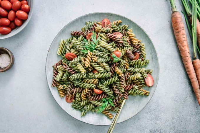 pesto pasta with cherry tomatoes on a white plate on a white table with carrots and a bowl of tomatoes