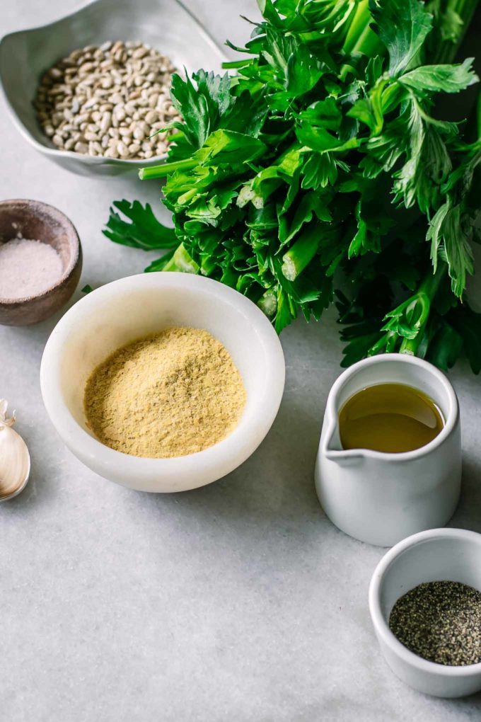 celery leaves, nuts, cheese, olive oil, salt, and pepper in small bowls on a marble countertop