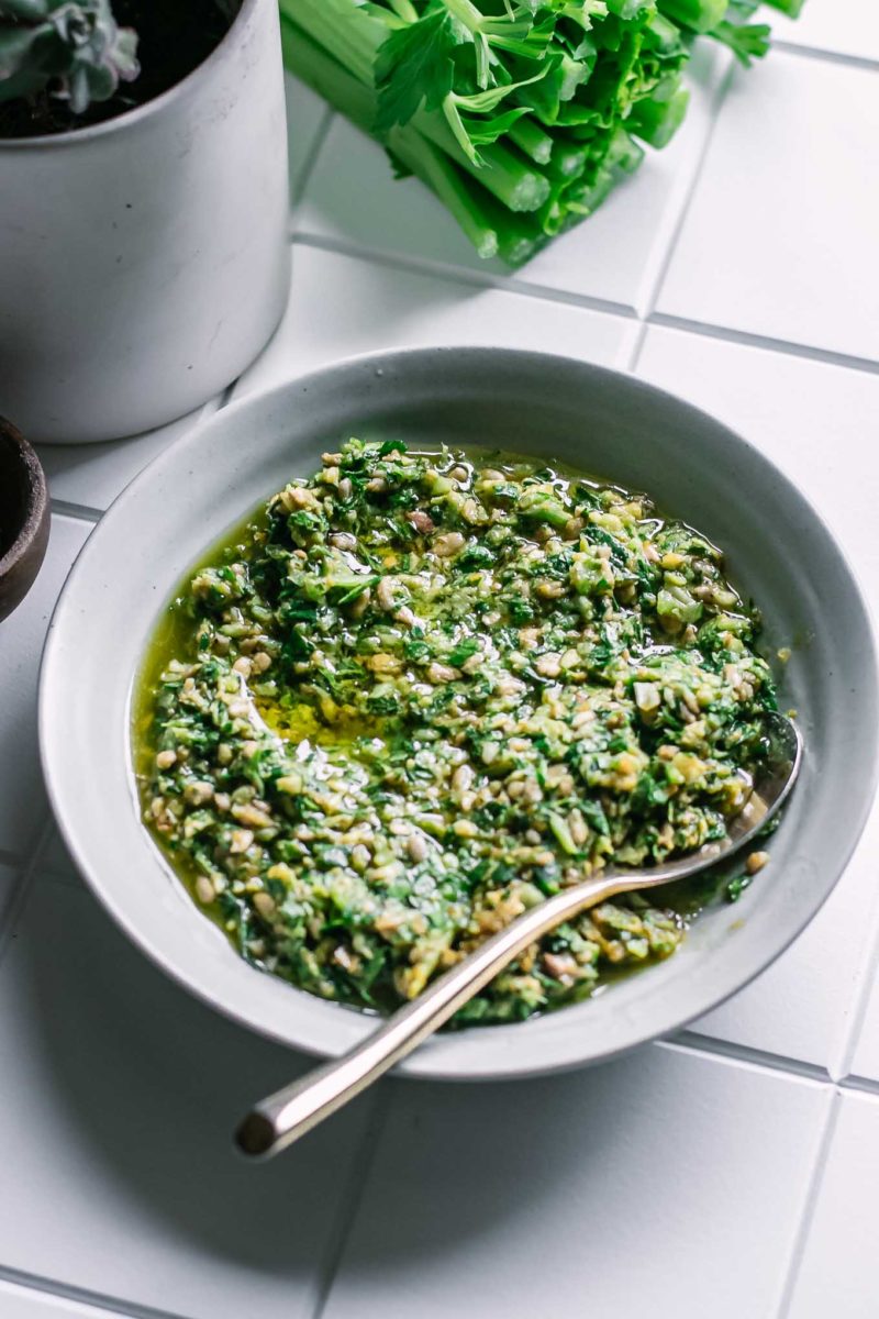 pesto with celery leaves in a small white bowl with a gold fork on a white table