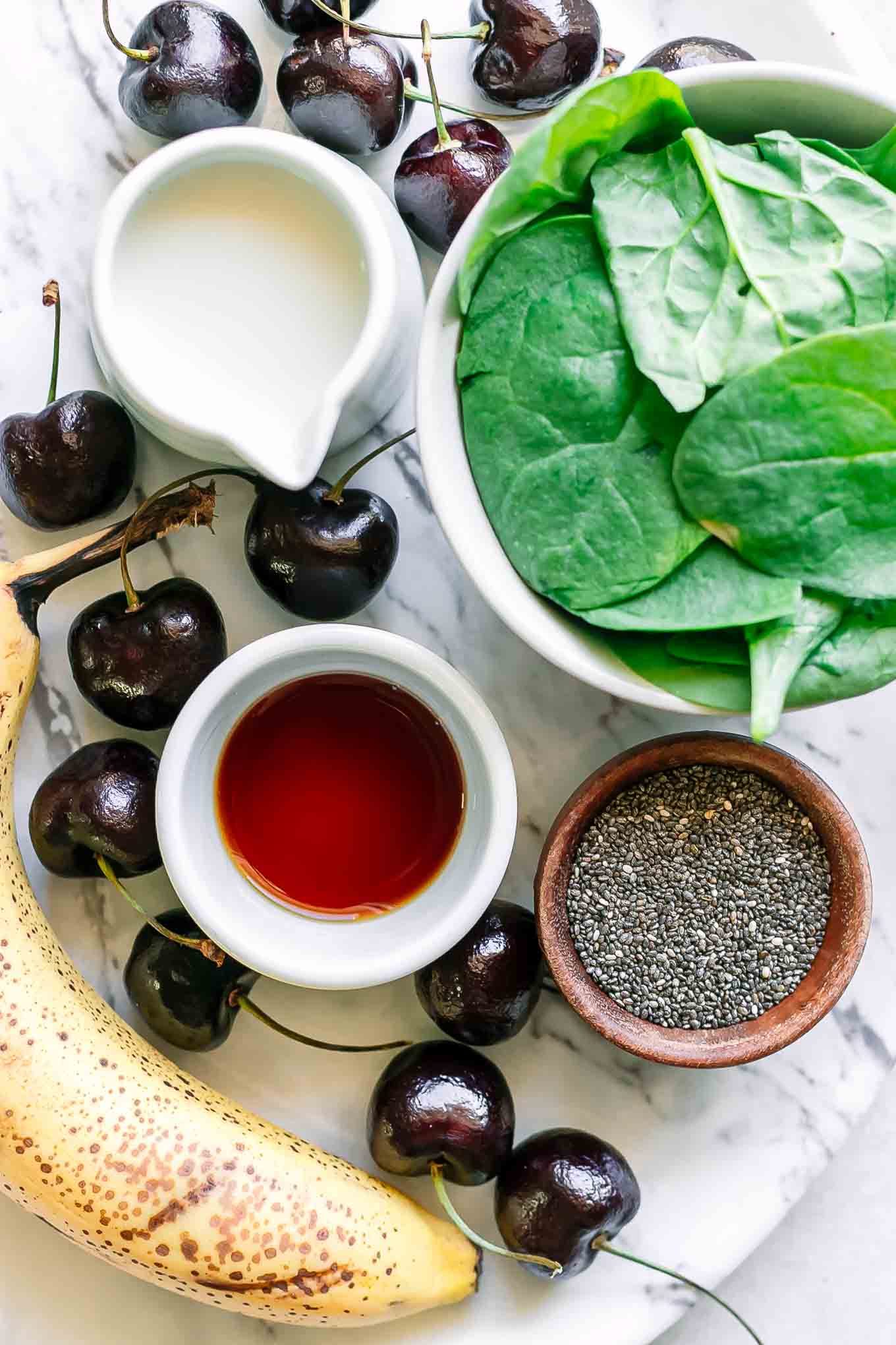 bowls of spinach, cherries, banana, chia seeds, milk, and vanilla on a table for a smoothie