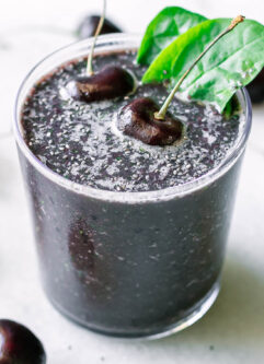 a dark purple cherry spinach smoothie in a glass with a cherry and spinach as garnish on a white table with fresh cherries
