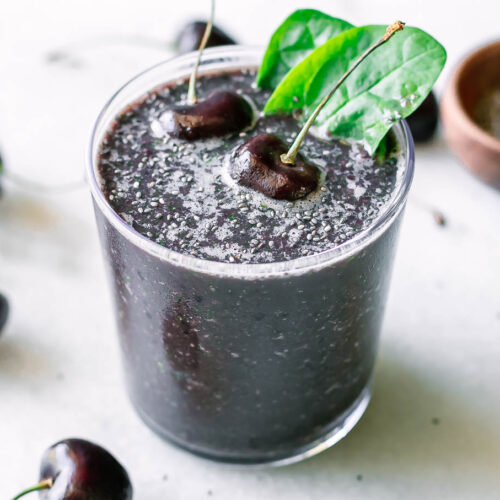 a dark purple cherry spinach smoothie in a glass with a cherry and spinach as garnish on a white table with fresh cherries