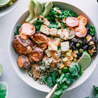 a white bowl with quinoa, black beans, tofu, sweet potatoes, and a creamy red chipotle chili lime sauce