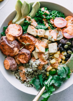 a white bowl with quinoa, black beans, tofu, sweet potatoes, and a creamy red chipotle chili lime sauce