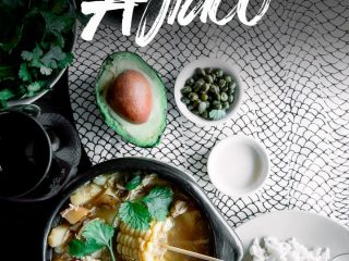 A chamba pot with Colombian ajiaco on a wood table with a side of rice, avocado, cream, and cilantro and a glass of wine and the words "Colombian Ajiaco"