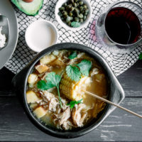 A chamba pot with Colombian ajiaco on a wood table with a side of rice, avocado, cream, and cilantro and a glass of wine.