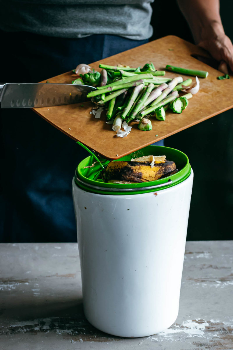 a woman scooping vegetables from a cutting board into a compost bin