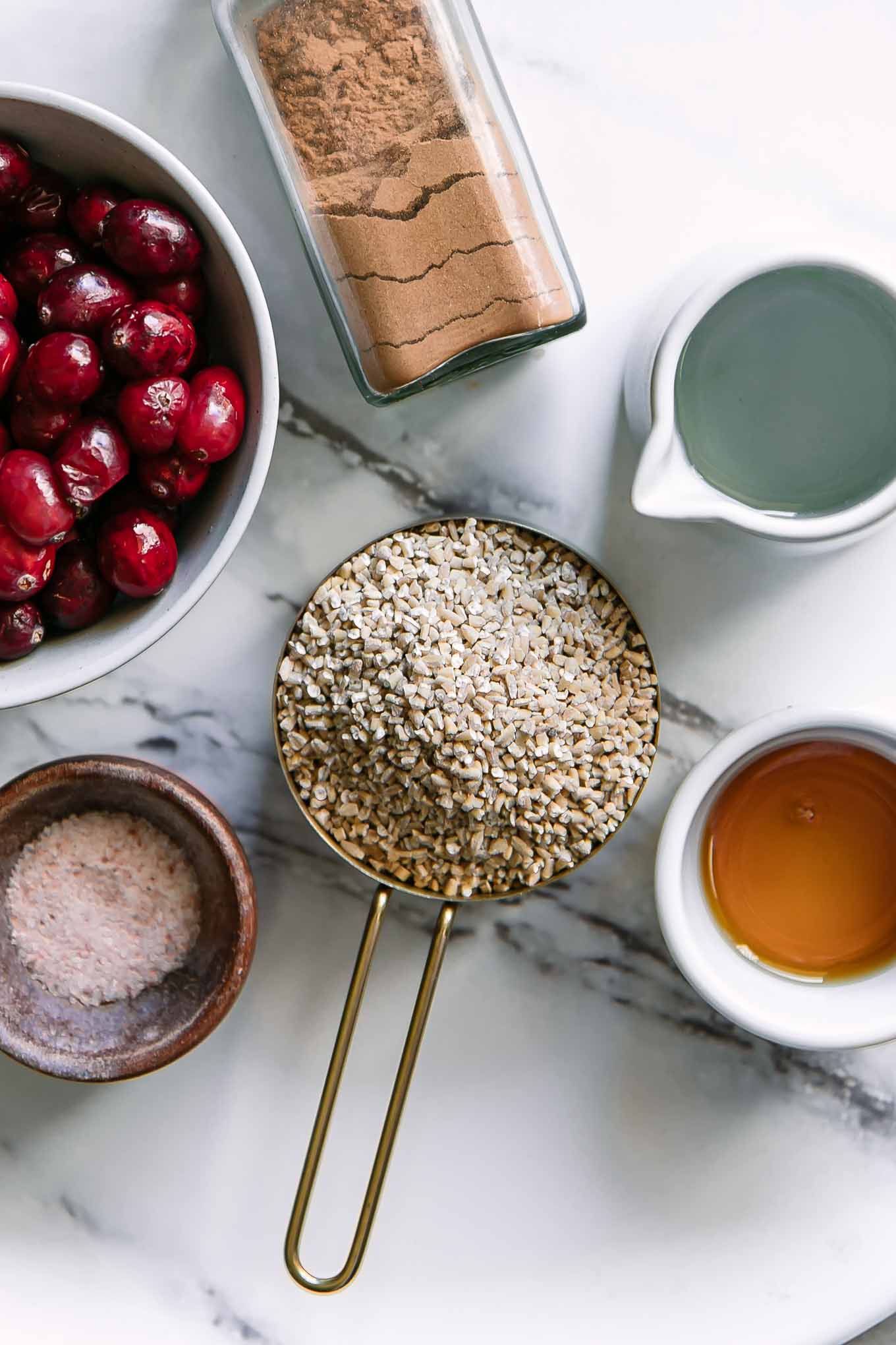a measuring cup with oatmeal, and bowls of cranberries, maple syrup, salt, water, cinnamon, and nutmeg for cranberry oatmeal