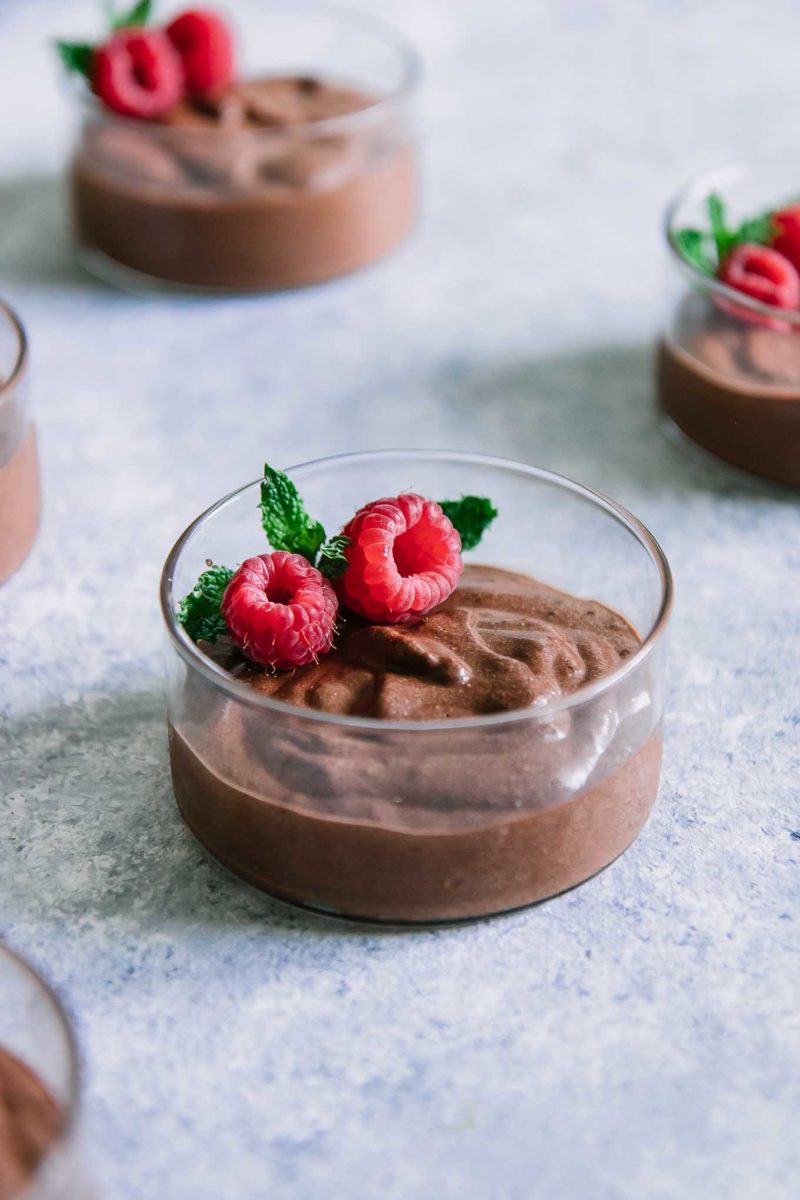 a glass bowl with brown chocolate mousse, red raspberries, and a sprig of green mint on a blue table