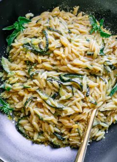 a black bowl with creamy zucchini orzotto pasta with a gold fork on a table with fresh zucchini