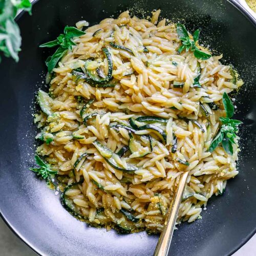 a black bowl with creamy zucchini orzotto pasta with a gold fork on a table with fresh zucchini