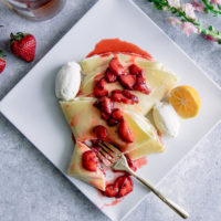 A crepe with ricotta and vanilla bourbon strawberries on a white plate on a blue table with flowers.