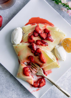 A crepe with ricotta and vanilla bourbon strawberries on a white plate on a blue table with flowers.