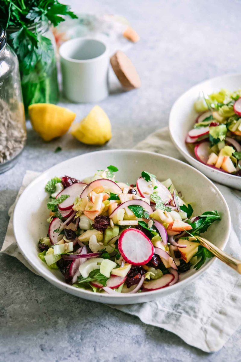 a colorful celery, apple, cranberry, and herb salad in a white bowl on a table with a white napkin