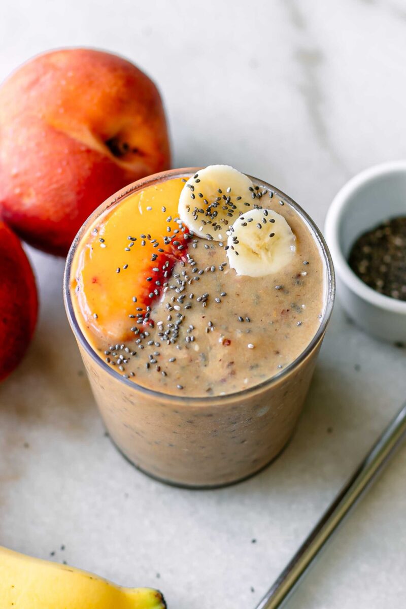 a peach and banana smoothie on a white table with whole peaches and a reusable straw