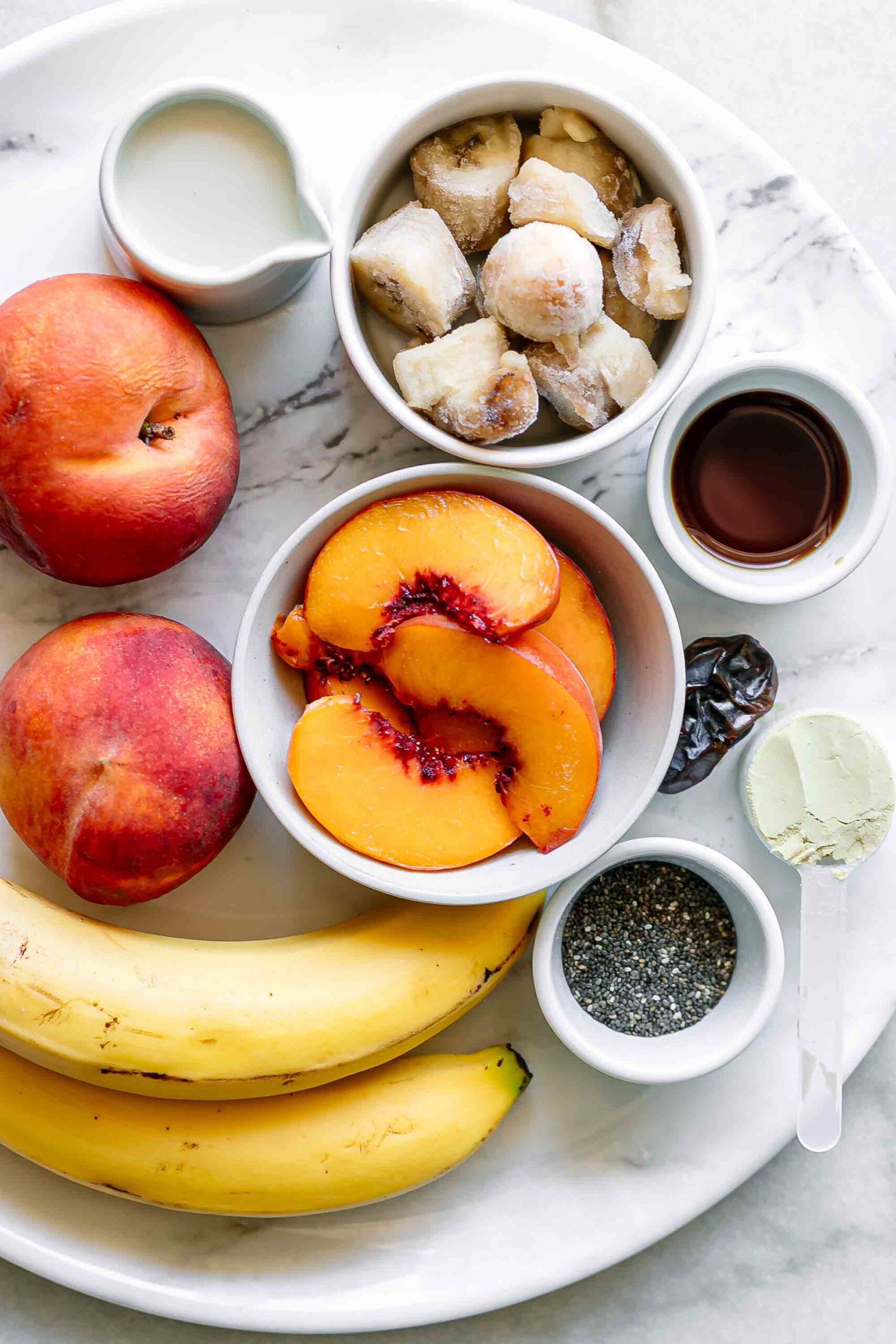 frozen peaches, frozen banans, chia seeds, vanilla, milk, a date, and protein powder on a white countertop for a smoothie