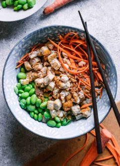 a rice and tofu bowl with edamame, carrots, and green onions on a white table