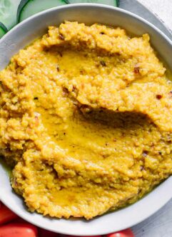 a bowl of orange-colored golden beet hummus dip with cut vegetables for dipping on a white table
