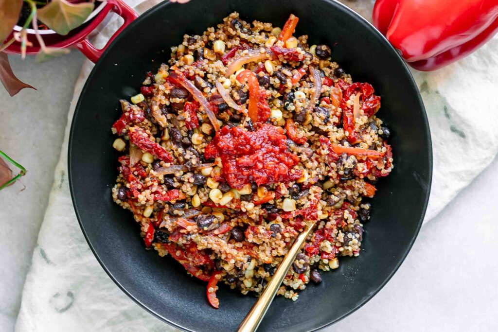 a quinoa bowl with black beans, bell peppers, onion, sun-dried tomatoes, and spicy harissa sauce on a white table with a gold fork