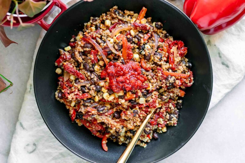 a quinoa bowl with black beans, bell peppers, onion, sun-dried tomatoes, and spicy harissa sauce on a white table with a gold fork