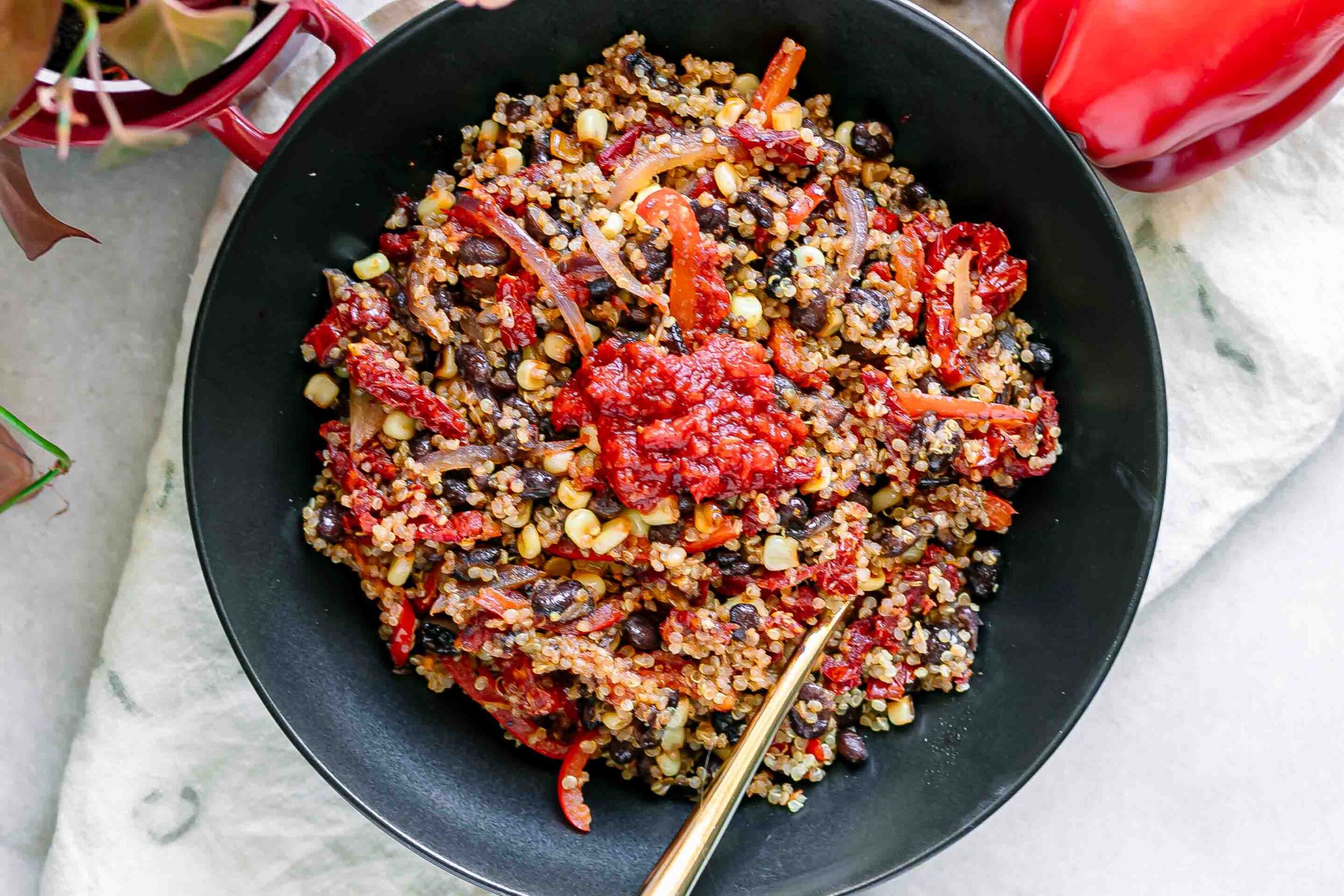 a quinoa bowl with black beans, bell peppers, onion, sun-dried tomatoes, and spicy harissa sauce on a white table with a gold fork