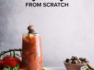 a bloody mary in a tall glass on a white table with tomatoes, basil, olives, and limes