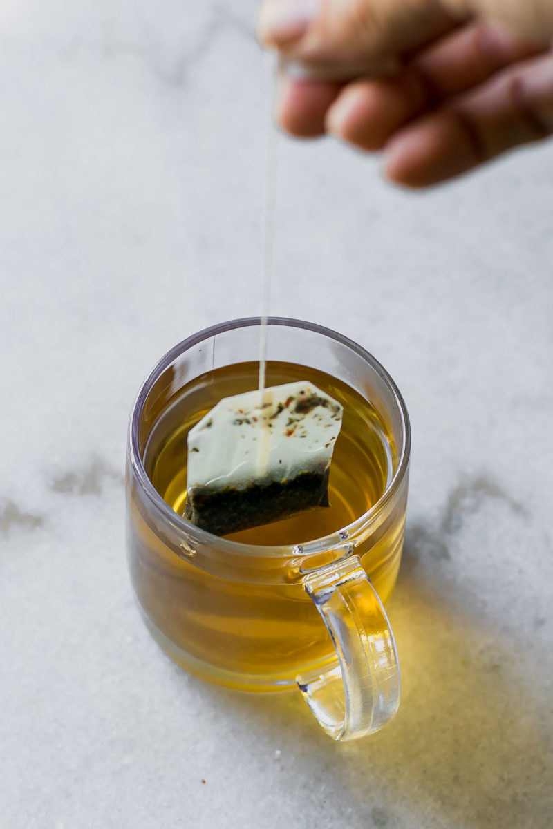 a hand pulling a tea bag out of a glass mug of brewed tea