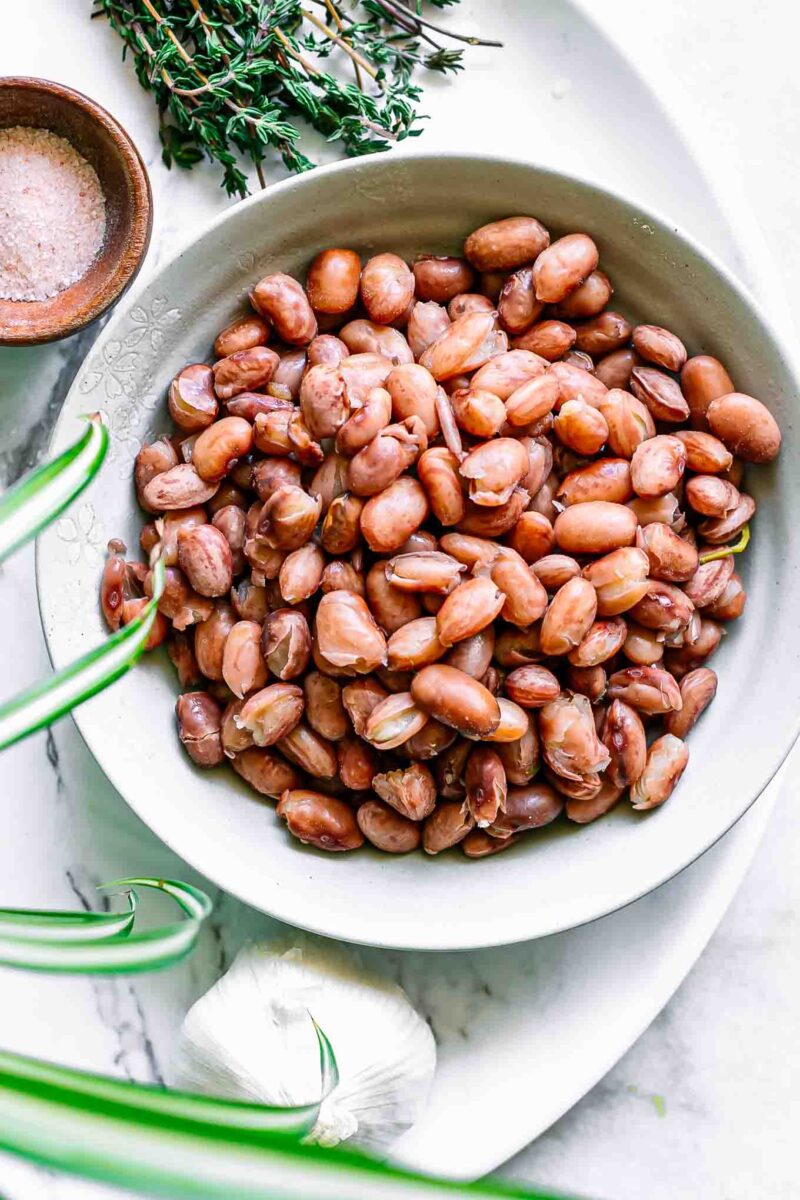 a bowl of cooked cranberry beans on a white table with herbs and spices